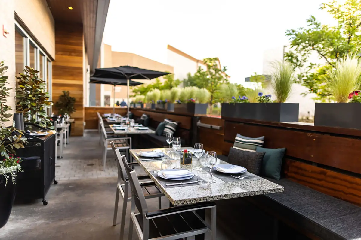 Knife Steakhouse patio in Plano — set tables under an umbrella, planters of grasses along the wood banquette
