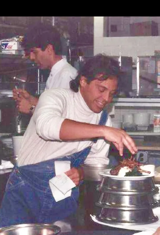 Archival photo of a young John Tesar plating in a kitchen alongside Anthony Bourdain